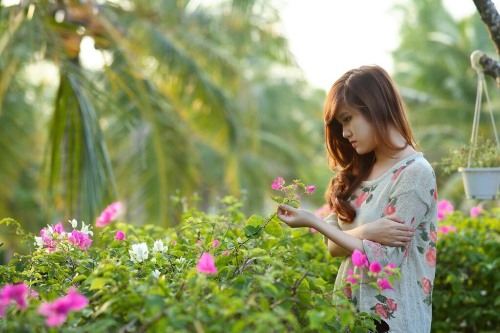Pensive young woman holding a flower, surrounded by lush garden flora.